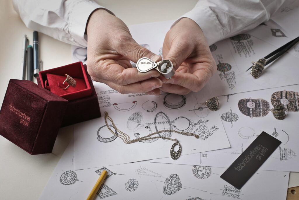 Close-up of hands inspecting a diamond ring with a loupe on a workbench filled with jewelry sketches.